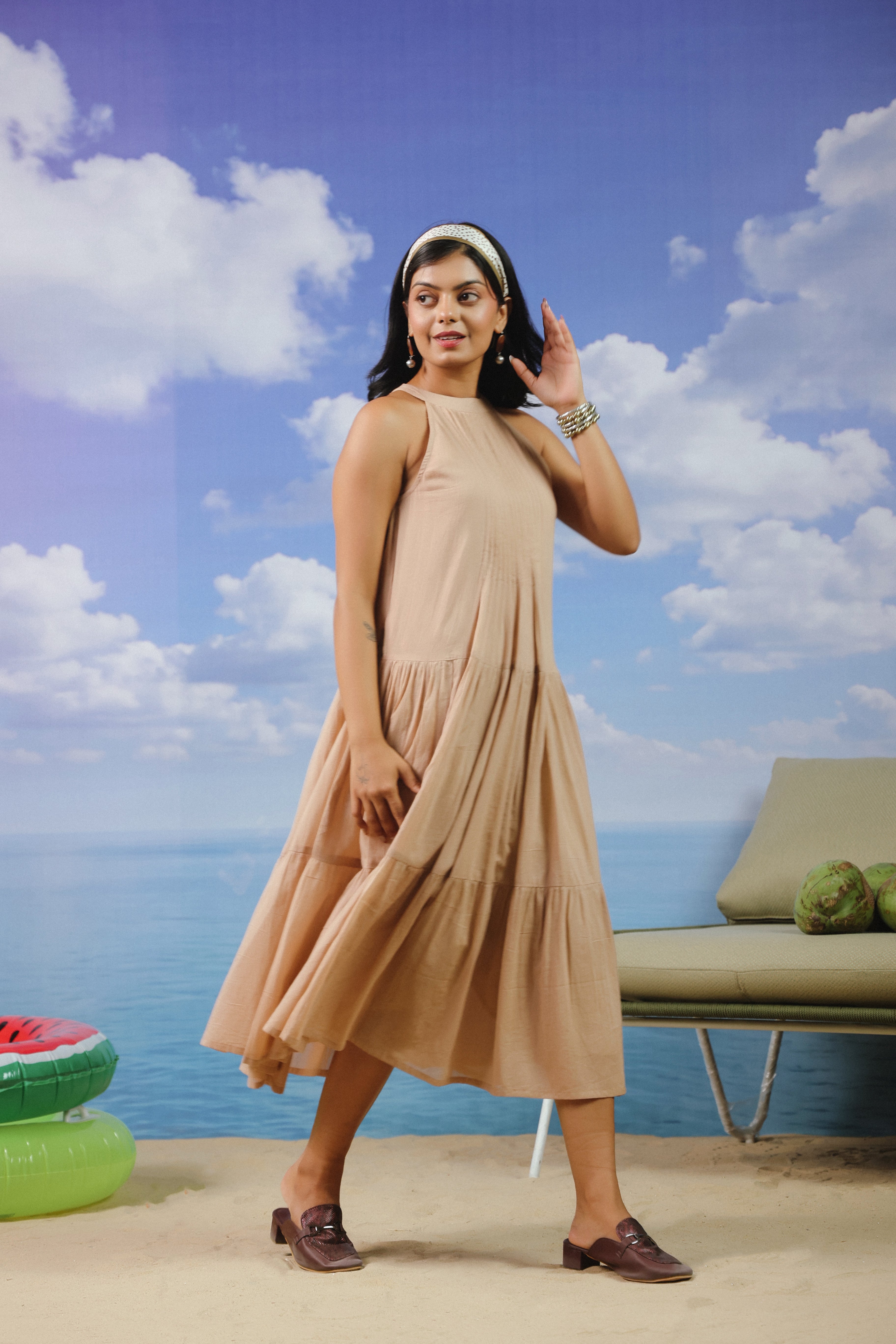 Woman wearing a beige sleeveless cotton midi dress with a high halter neckline, pleated bodice, and flowy tiered skirt, offering a chic and elegant look perfect for vacations, resort wear, beach outings, brunches, and summer occasions.