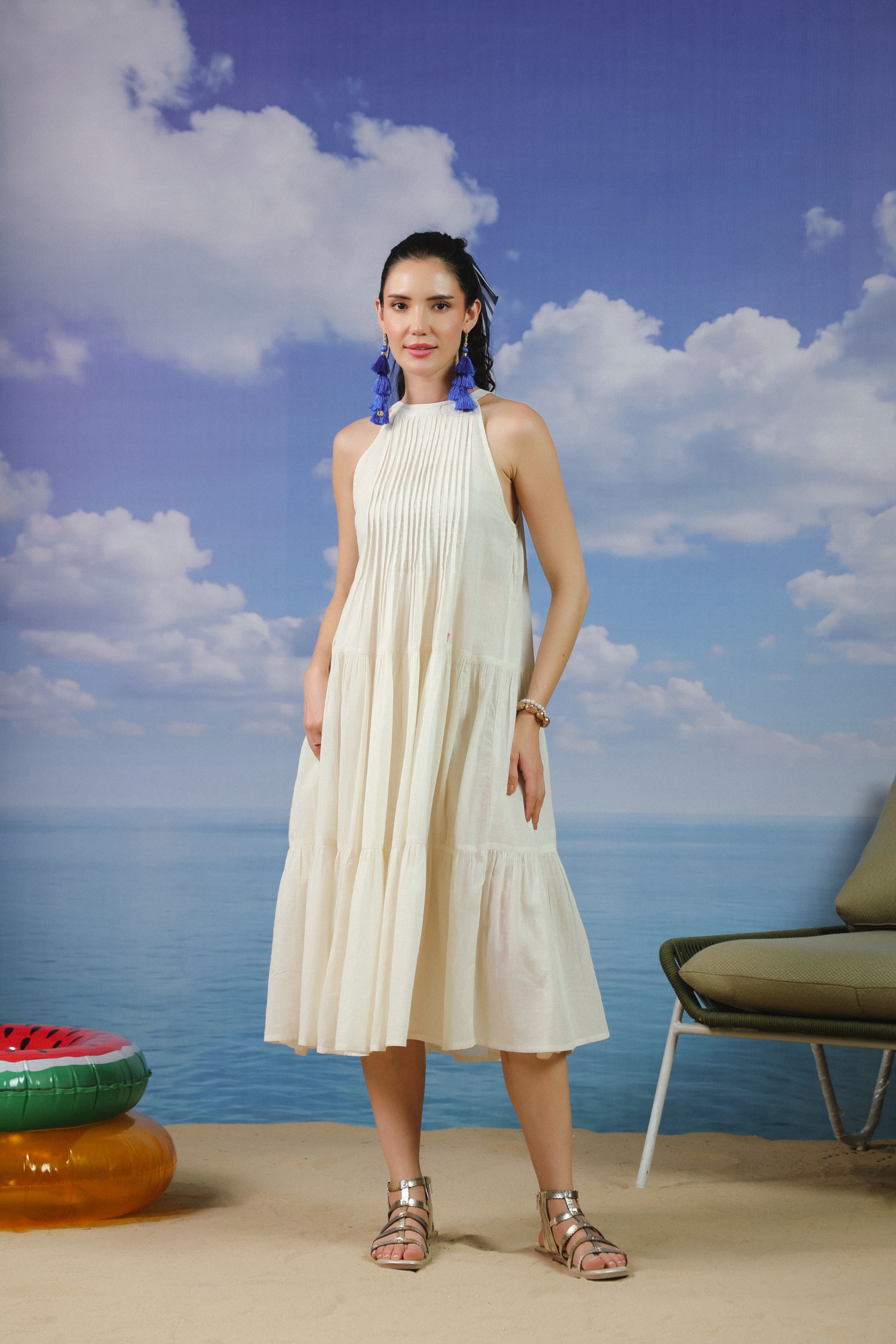 Woman wearing an ivory sleeveless cotton midi dress with a high halter neckline, elegant pleated bodice, and flowy tiered skirt, creating a timeless and sophisticated look for vacations, beach outings, brunches, resort wear, and summer occasions.