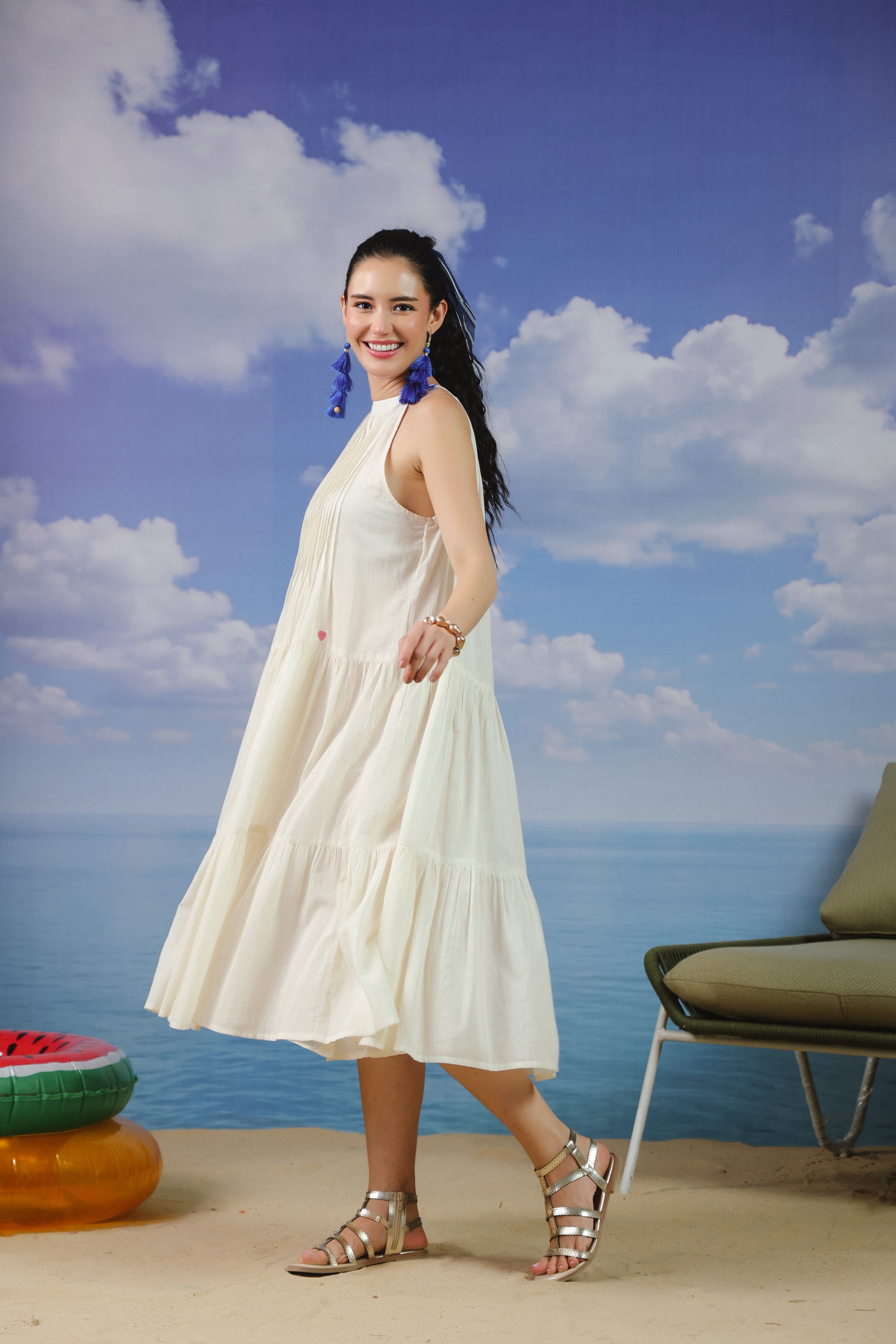 Woman wearing an ivory sleeveless cotton midi dress with a high halter neckline, elegant pleated bodice, and flowy tiered skirt, creating a timeless and sophisticated look for vacations, beach outings, brunches, resort wear, and summer occasions.