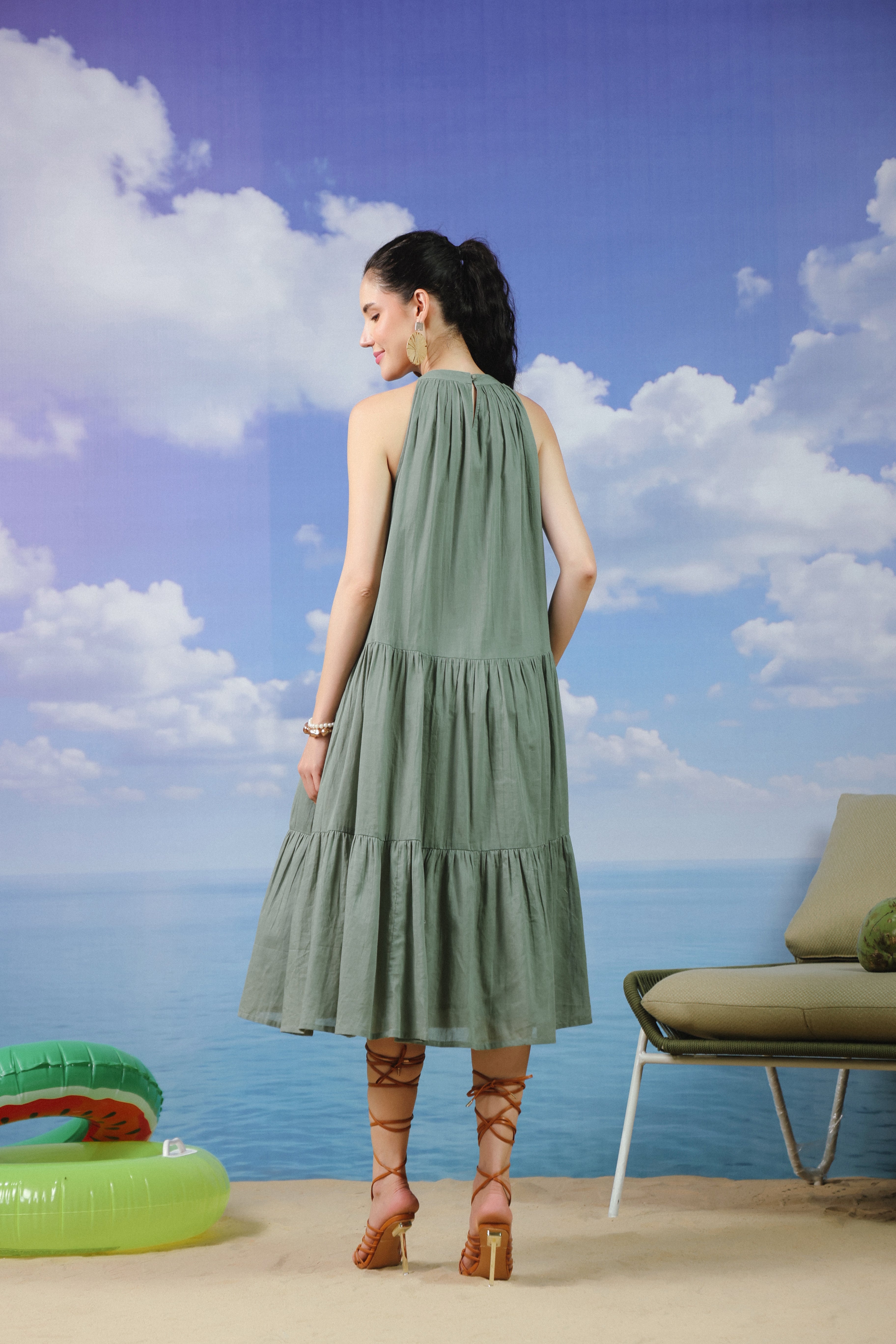 Woman wearing a sage green sleeveless cotton midi dress with a high halter neckline, pleated bodice, and tiered flared skirt, creating an elegant and flowy look ideal for summer vacations, beach outings, brunches, and resort wear.
