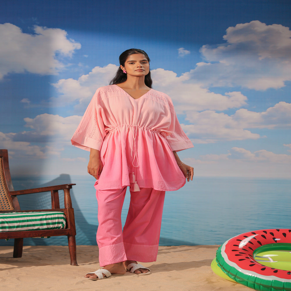 Woman wearing a soft pink cotton co-ord set featuring a relaxed drawstring tunic with lace detailing and matching straight-leg pants, styled with white sandals on a beach-themed backdrop