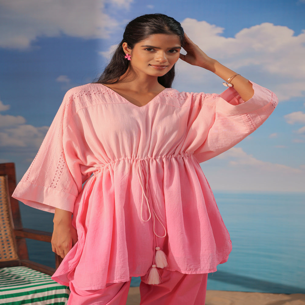 Woman wearing a soft pink cotton co-ord set featuring a relaxed drawstring tunic with lace detailing and matching straight-leg pants, styled with white sandals on a beach-themed backdrop