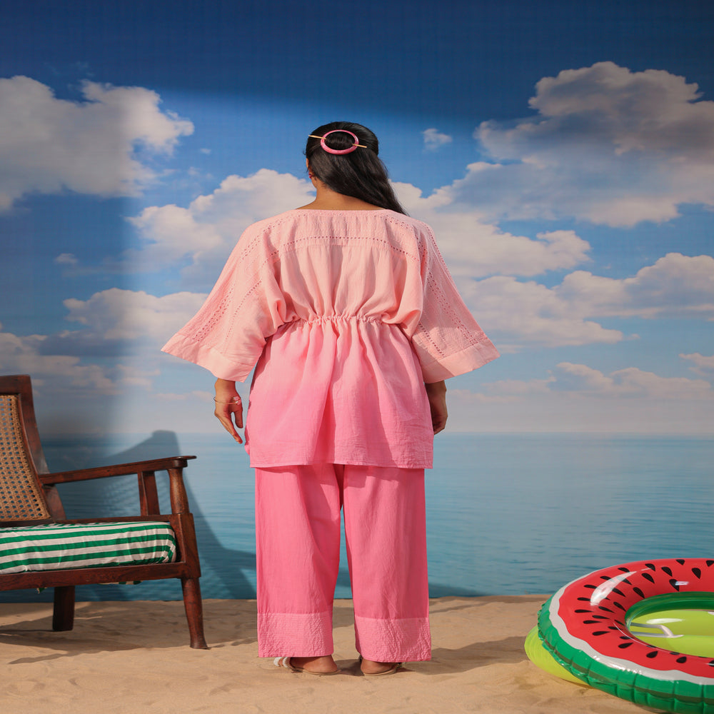 Woman wearing a soft pink cotton co-ord set featuring a relaxed drawstring tunic with lace detailing and matching straight-leg pants, styled with white sandals on a beach-themed backdrop