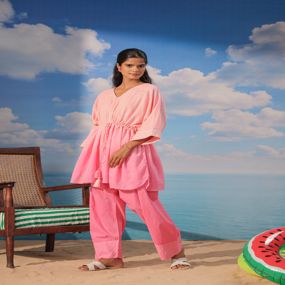 Woman wearing a soft pink cotton co-ord set featuring a relaxed drawstring tunic with lace detailing and matching straight-leg pants, styled with white sandals on a beach-themed backdrop