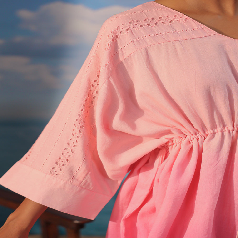 Woman wearing a soft pink cotton co-ord set featuring a relaxed drawstring tunic with lace detailing and matching straight-leg pants, styled with white sandals on a beach-themed backdrop