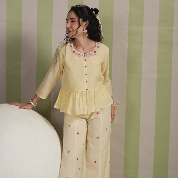 Woman wearing a blush yellow embroidered peplum co-ord set with wide-leg palazzo pants, featuring a floral embroidered neckline and relaxed elegant fit. Perfect for casual and festive occasions.