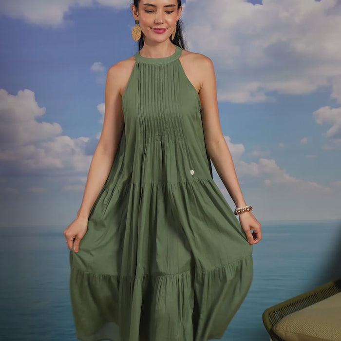 Woman wearing a sage green sleeveless cotton midi dress with a high halter neckline, pleated bodice, and tiered flared skirt, creating an elegant and flowy look ideal for summer vacations, beach outings, brunches, and resort wear.