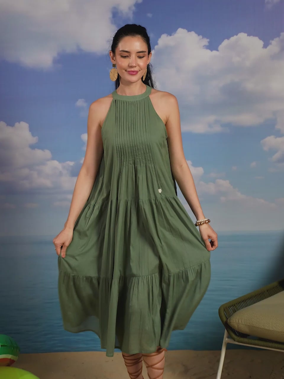 Woman wearing a sage green sleeveless cotton midi dress with a high halter neckline, pleated bodice, and tiered flared skirt, creating an elegant and flowy look ideal for summer vacations, beach outings, brunches, and resort wear.